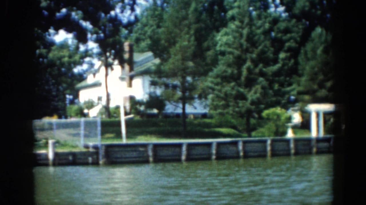 Waterfront residential property seen from the river, with a white house and ornamental garden structure amid mature summer trees.