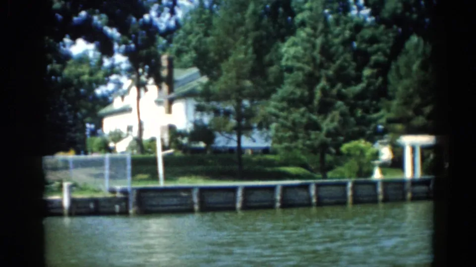 1962 Illinois riverfront home with white siding and ornamental garden structure visible through dense summer foliage, on 8mm film.