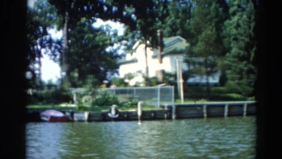 1962 Illinois river panning across waterfront homes, a red boat moored near a wooden dock, and dense green summer foliage, on 8mm film.