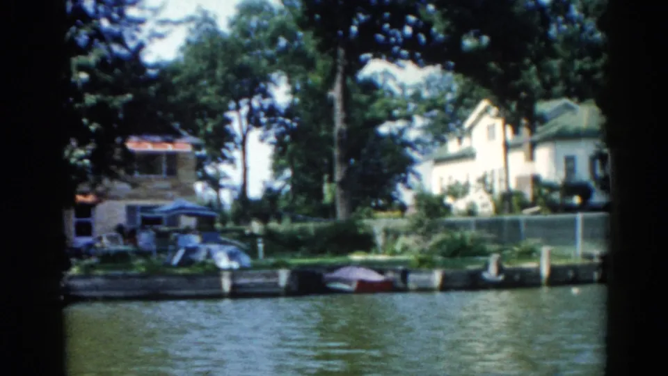 1962 Illinois riverfront with a red-roofed boathouse and multi-storey house visible through summer trees, red boat near the bank, on 8mm film.