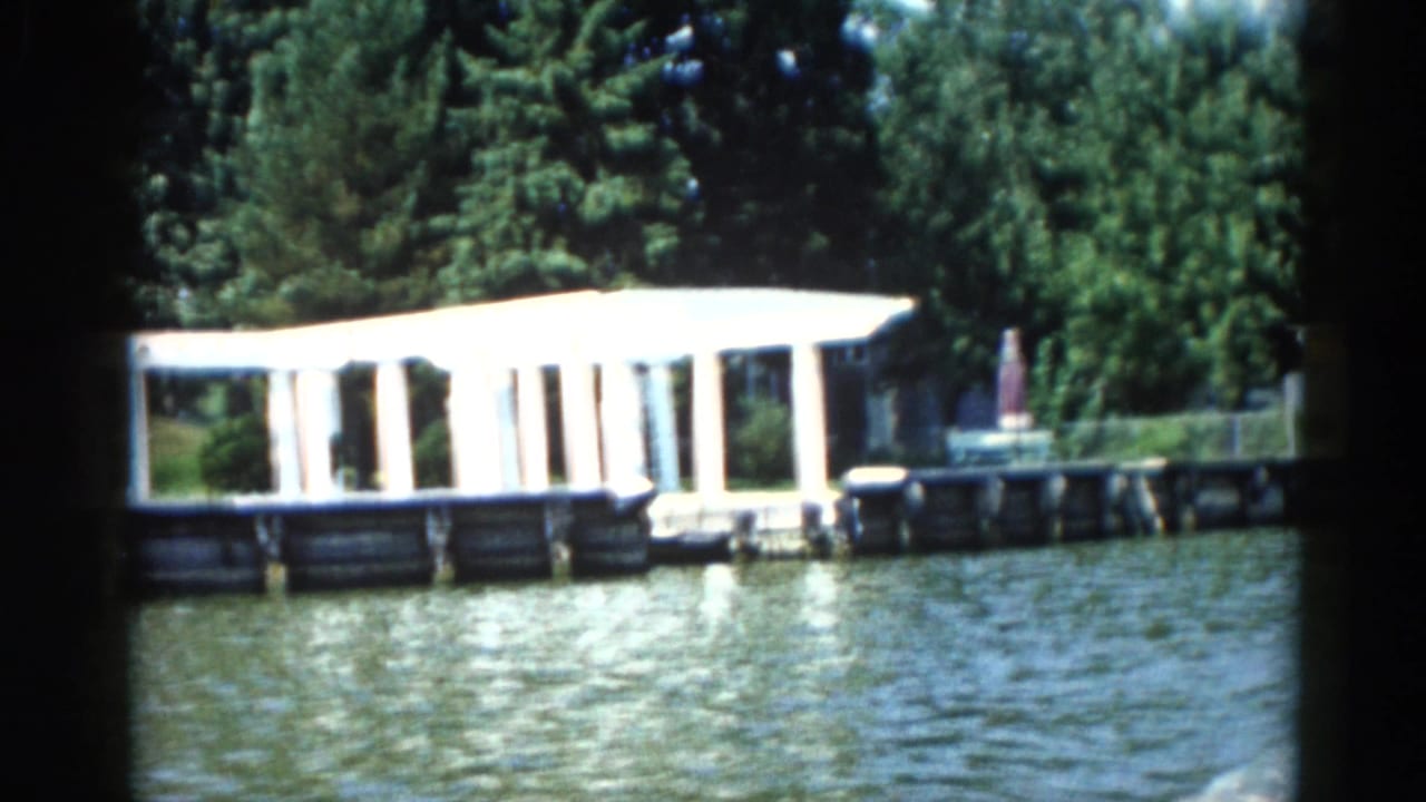 1962 8mm home movie frame of a white colonnaded dock pavilion on an Illinois lake, with summer tree cover behind and rippled water in the foreground.