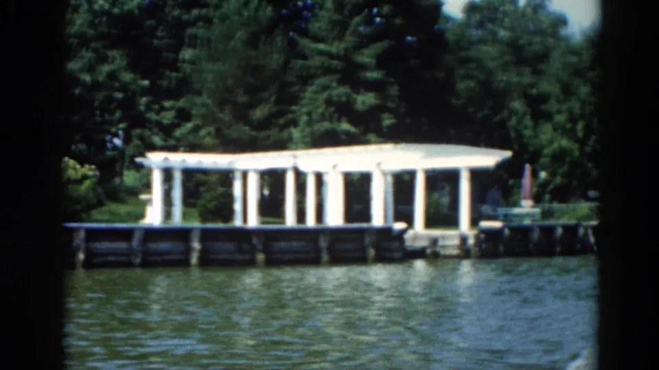 1962 8mm home movie frame showing a white colonnaded dock pavilion on an Illinois lake, with dense deciduous trees and a small red accent object in view.