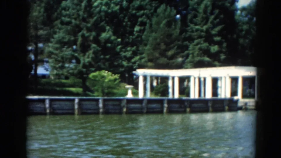 1962 8mm home movie wide shot of a long white colonnaded dock structure along an Illinois lakeshore, with full summer tree canopy behind.