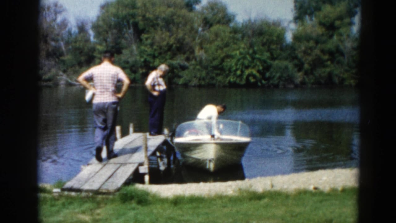Two adults on a wooden dock at an Illinois lake, circa 1962, with a small motorboat moored alongside; a third person leans into the vessel.