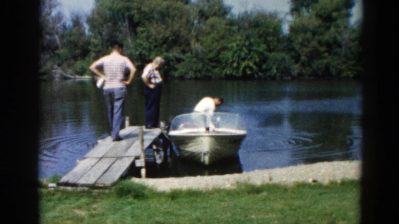 Continued wide shot of three figures at a wooden Illinois lakeside dock, circa 1962, with a small motorboat moored alongside and dense summer foliage.