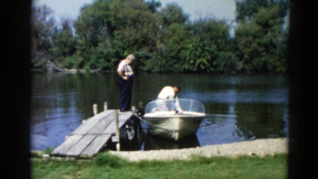 Two men on a wooden dock with a small motorboat moored at a gravel shoreline, calm lake and trees in background.
