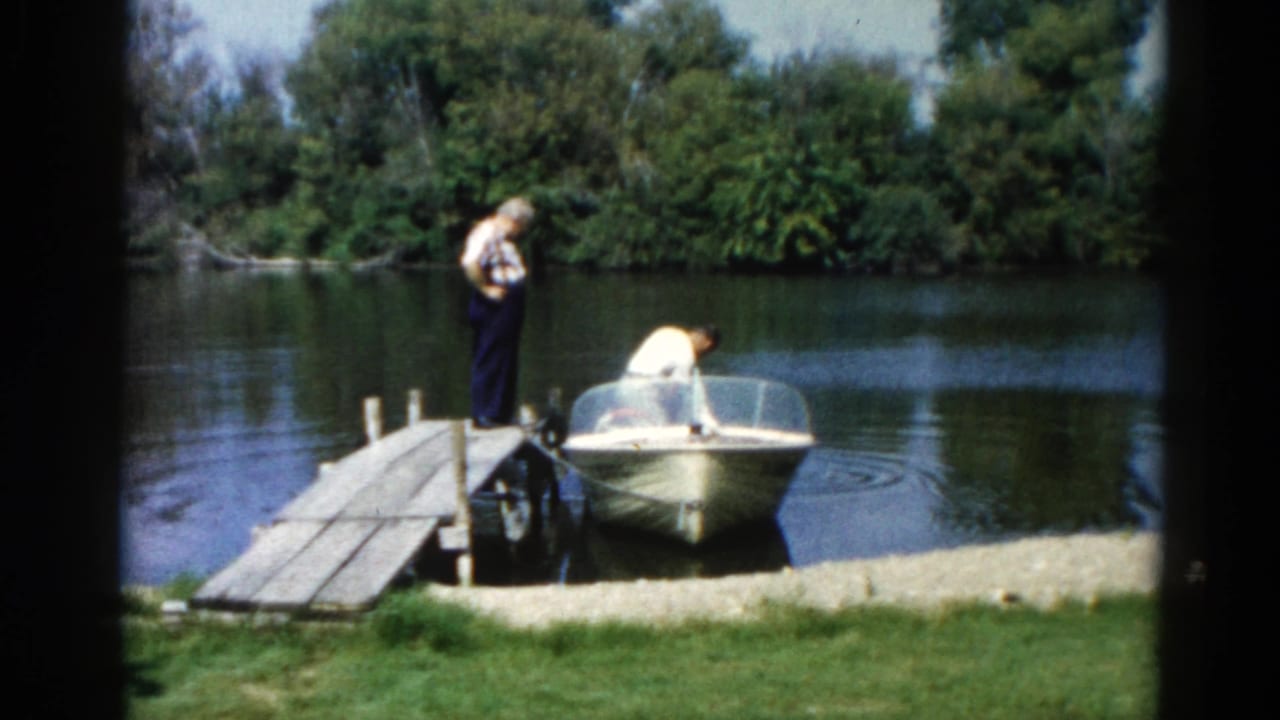 Two men on a wooden dock beside a small motorboat, calm water and wooded bank behind them.