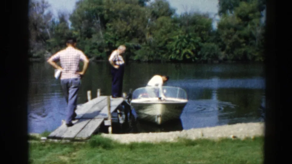 Three men at a lake dock in casual summer dress, one watching with hands on hips as another tends to a motorboat, Illinois, circa 1962.