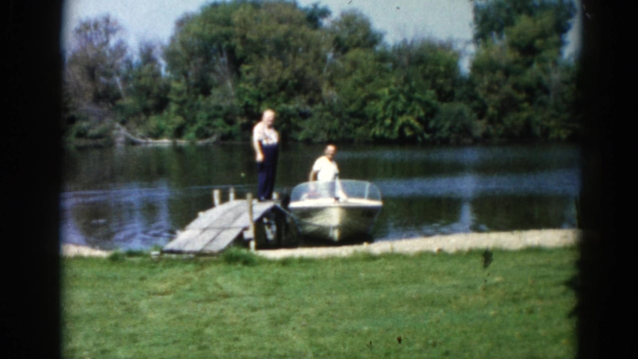 Two men at a wooden dock preparing a small motorboat on a calm lake, green lawn and treeline behind.