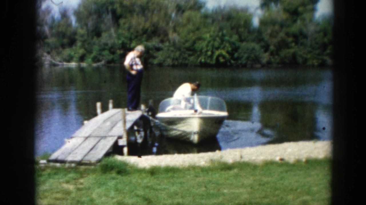 Two men at a wooden dock, one leaning into a motorboat, calm lake and trees behind.