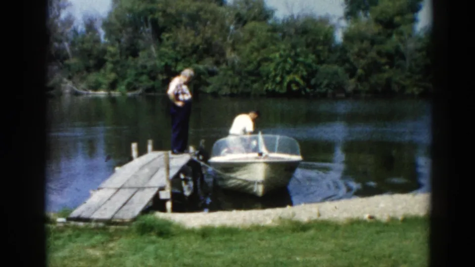Two men on a wooden dock attending to a mid-century runabout; plaid shirt and white shirt visible, Illinois lakeside 1962 8mm.
