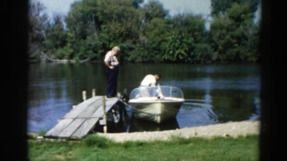 Man works on outboard motor while companion watches on a wooden dock, Illinois lake, 1962 8mm home movie.