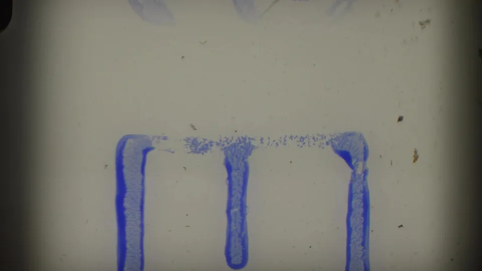 Film leader or damaged 8mm frame from the start of the reel, showing blue ink markings on a gray film base with surface scratches.