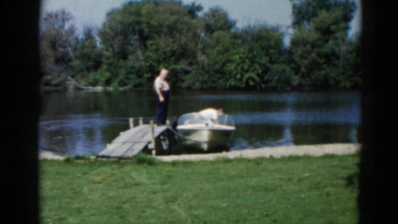 Wide establishing shot of a man at a wooden lakeside dock with a white motorboat moored alongside; Illinois lake, circa 1962, 8mm color.