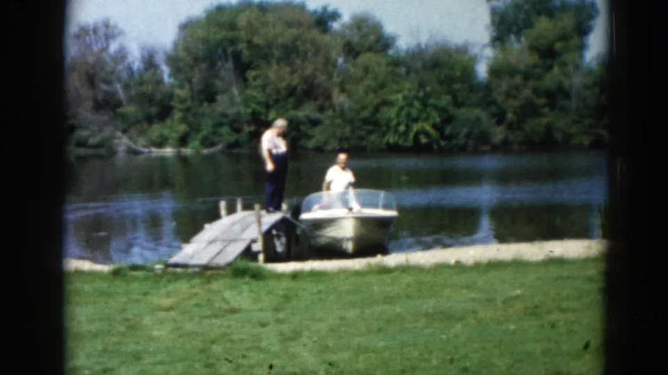 Two adult men interacting at a lakeside wooden dock beside a white motorboat; Illinois, circa 1962, captured on 8mm color film.
