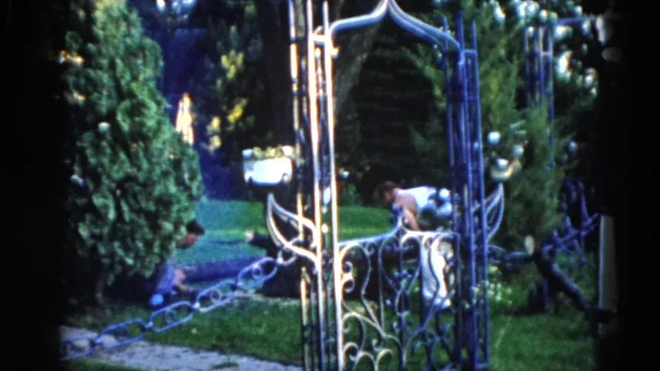 Ornate wrought-iron gate with a distant adult figure standing on a green lawn, from a 1962 Illinois 8mm backyard sequence.