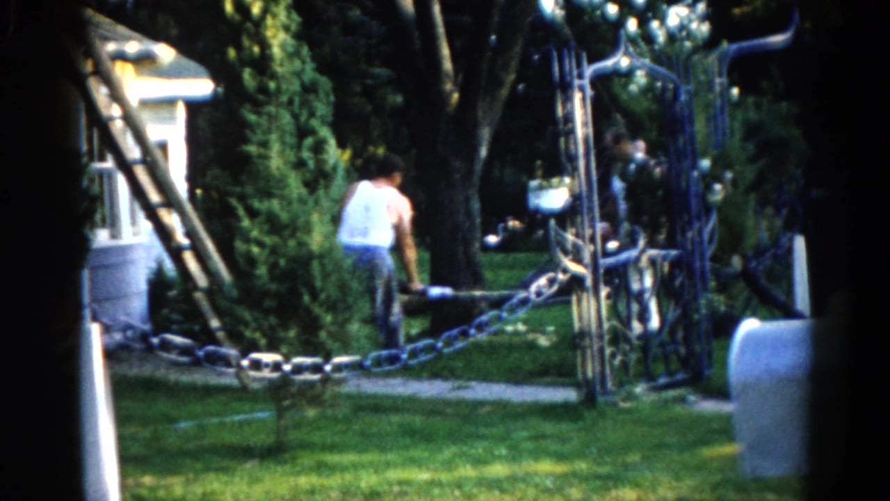 Two men working near a ladder in a suburban yard, c. 1962 Illinois — ornate wrought-iron gate and chain in foreground, white house and trees behind.
