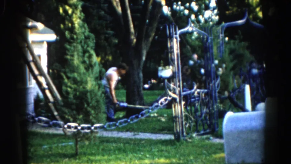 Man working at the base of a tree, c. 1962 Illinois — ornate wrought-iron gate and chain in foreground, ladder against tree, white house behind.
