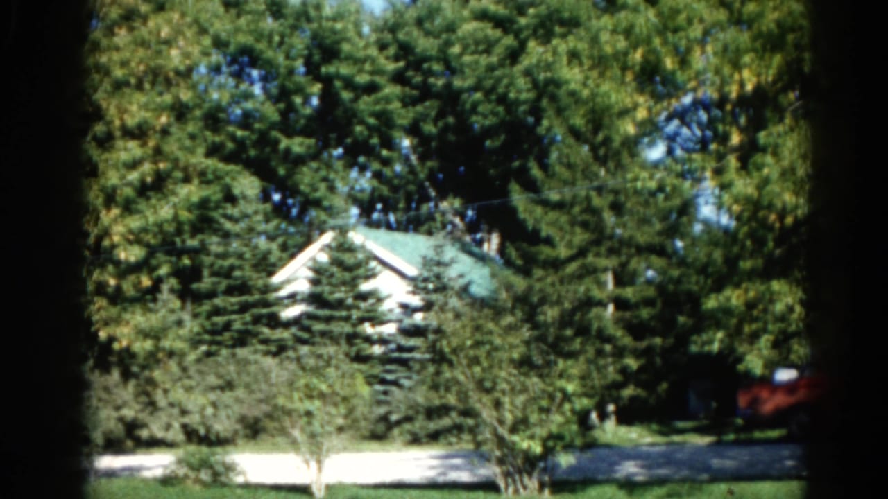 1962 Illinois home movie opening frame: white house with green roof partially obscured by a mature tree canopy; mowed lawn and light driveway below.