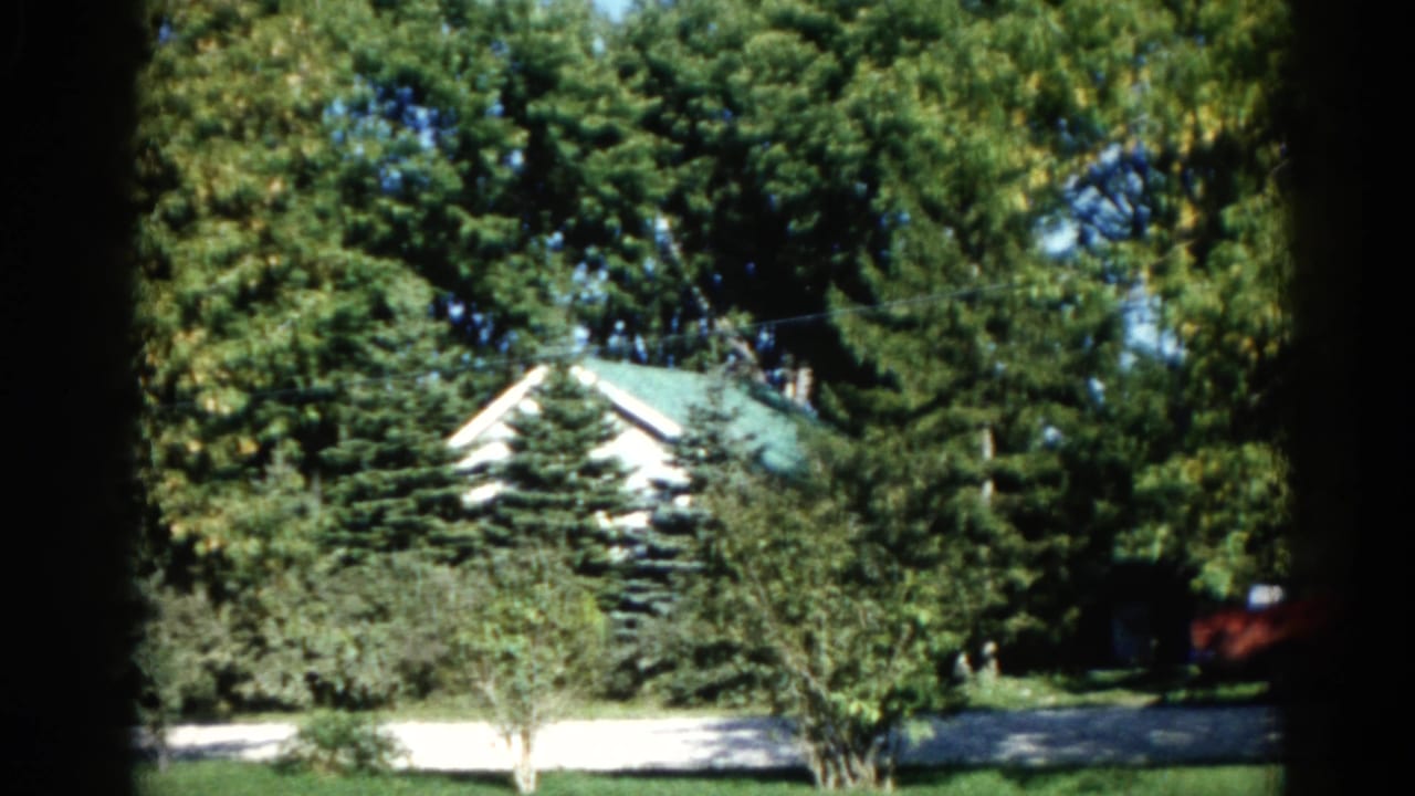White house with green roof seen through dense tree canopy; red outbuilding visible at far right, green lawn and driveway below.