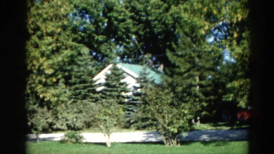 Illinois home movie frame circa 1962: white house with green roof partially obscured by a tall conifer; mowed lawn and driveway below, red structure at right.