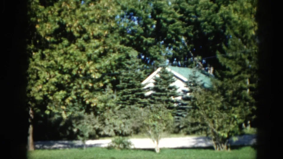 Final frame of 1962 Illinois home movie: green-roofed white house through summer tree canopy, mowed lawn and driveway below, red outbuilding at right.