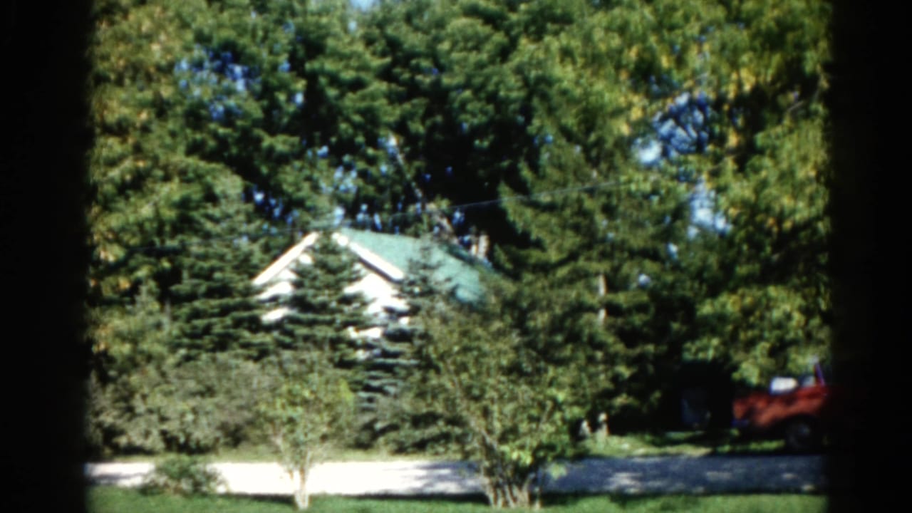 White house with green peaked roof seen through dense summer trees; red vehicle at right on driveway.