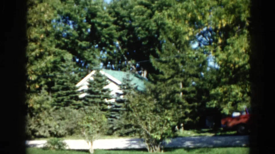 8mm home movie, Illinois 1962: mature tree canopy frames white house with green roof; red vehicle and driveway visible at right.