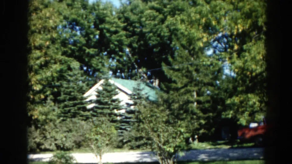 8mm home movie, Illinois 1962: white house with green peaked roof behind mature deciduous trees; red vehicle at right edge.