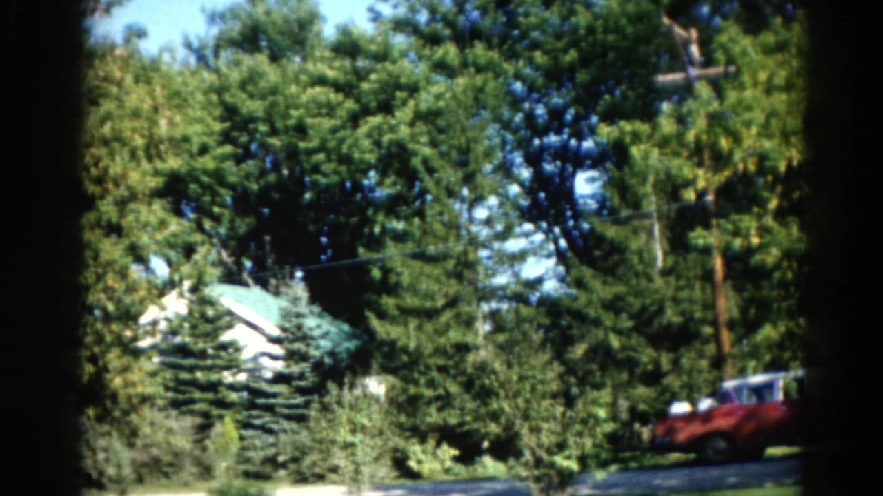 1962 Illinois home movie: white house with green roof partially concealed by mature deciduous trees, red vintage automobile at roadside, blue sky above.