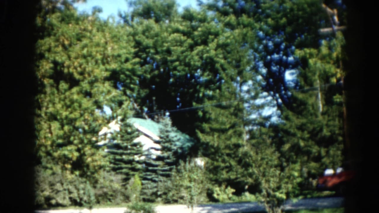 1962 Illinois home movie: suburban house with green roof behind full-leaf trees, red car at roadside, telephone pole visible above foliage.
