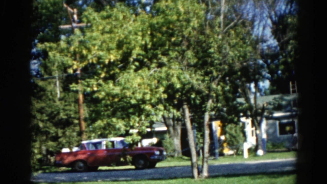 Dark red early-1960s American car on a tree-lined Illinois suburban street in summer, figure in white near roadside and utility pole visible, circa 1962.