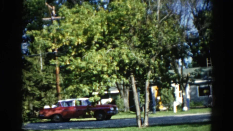 Dark red early-1960s American car on a tree-lined Illinois suburban street in summer, figure in white near roadside and utility pole visible, circa 1962.