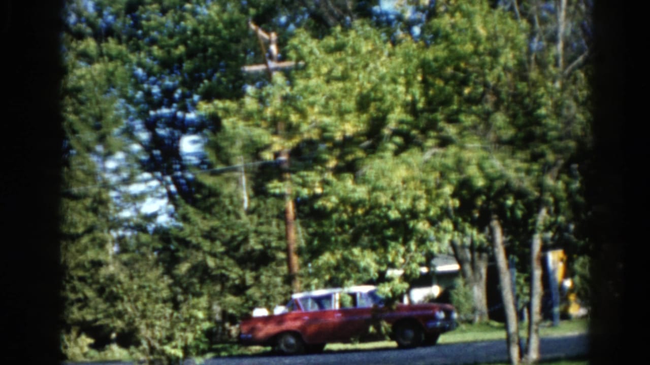 Dark red early-1960s American car mid-pan along an Illinois suburban street in summer, wooden utility pole and power lines prominent, circa 1962.