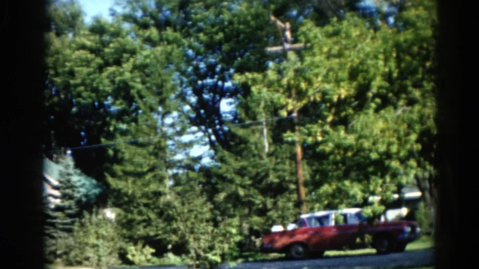 Camera pan reveals a suburban house with a green roof and mature trees in an Illinois neighborhood, red classic car at lower right, circa 1962.
