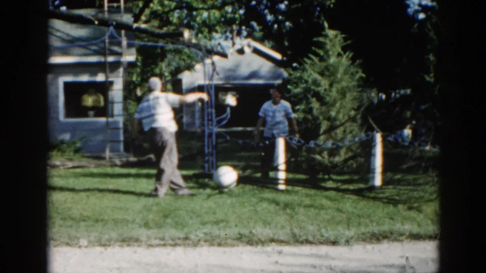 Two men collaborating at a pulley apparatus in a suburban Illinois backyard, circa 1962; one in striped shirt, one in light shirt, 8mm color film.