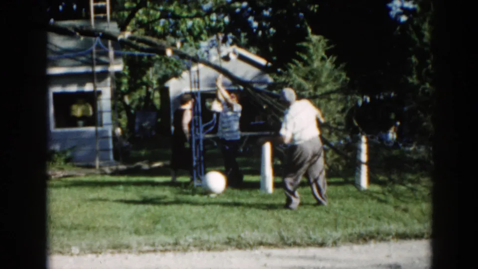 Two men in casual 1960s work clothing manage a rope-secured leaning tree in a suburban Illinois backyard, circa 1962, captured on 8mm film.