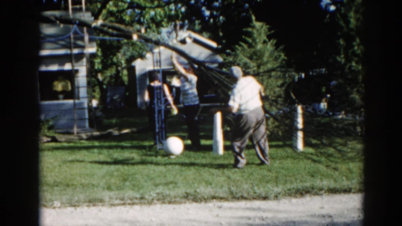 Two men beside a rope-secured tree structure in a suburban backyard; one in a light shirt, one in a striped shirt, white ball on grass.