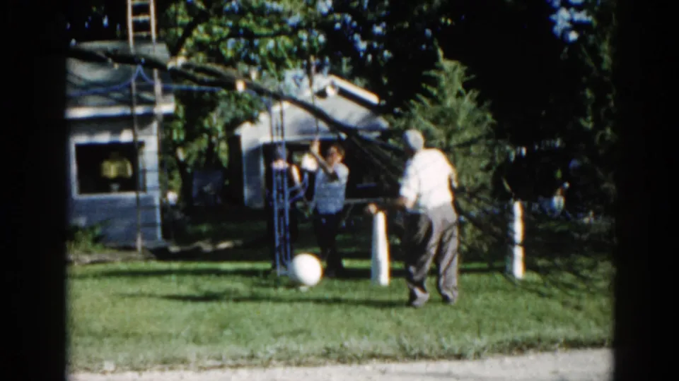 Man in striped shirt reaches up toward the tree as his companion stands nearby in a suburban Illinois backyard, circa 1962, on 8mm film.