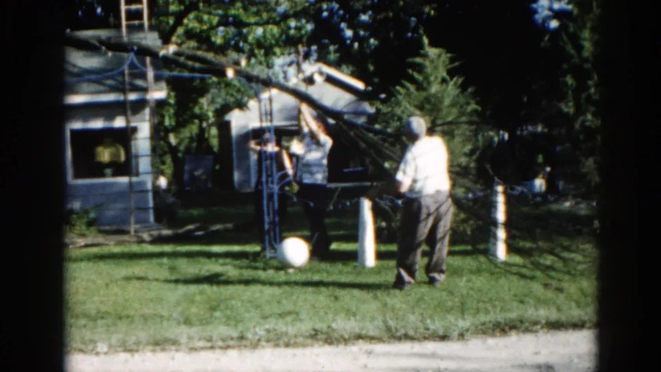Two men beside a rope-secured tree in a 1960s suburban Illinois backyard; period-typical residential house and hedges are visible, circa 1962.
