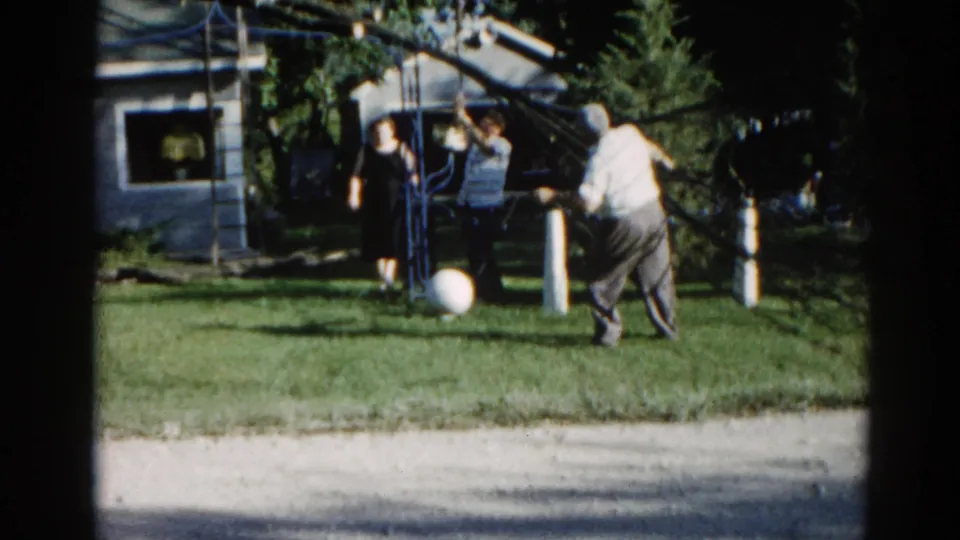 Two adults near a backyard tree, c.1962 suburban Illinois; white ball on green lawn and residential house visible behind foliage.