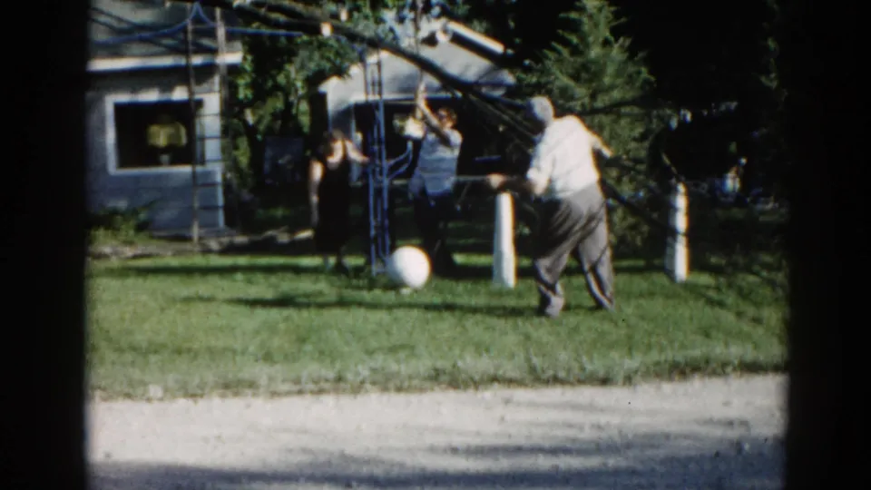 Adult and possible minor near tree during trimming activity, c.1962 suburban backyard; metal frame structure and white ball visible on lawn.