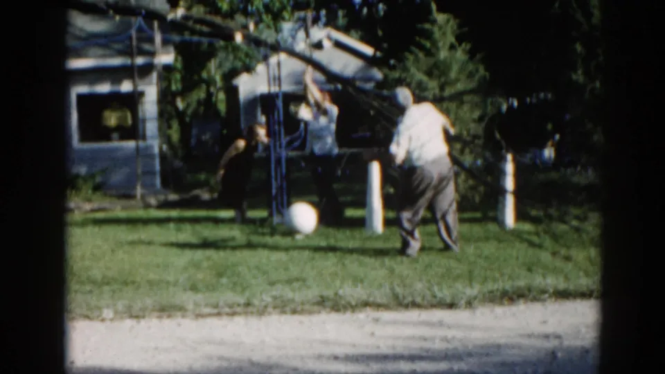 Man pauses near a tree in suburban backyard, c.1962 Illinois; white ball, white fence posts, and residential house visible in background.