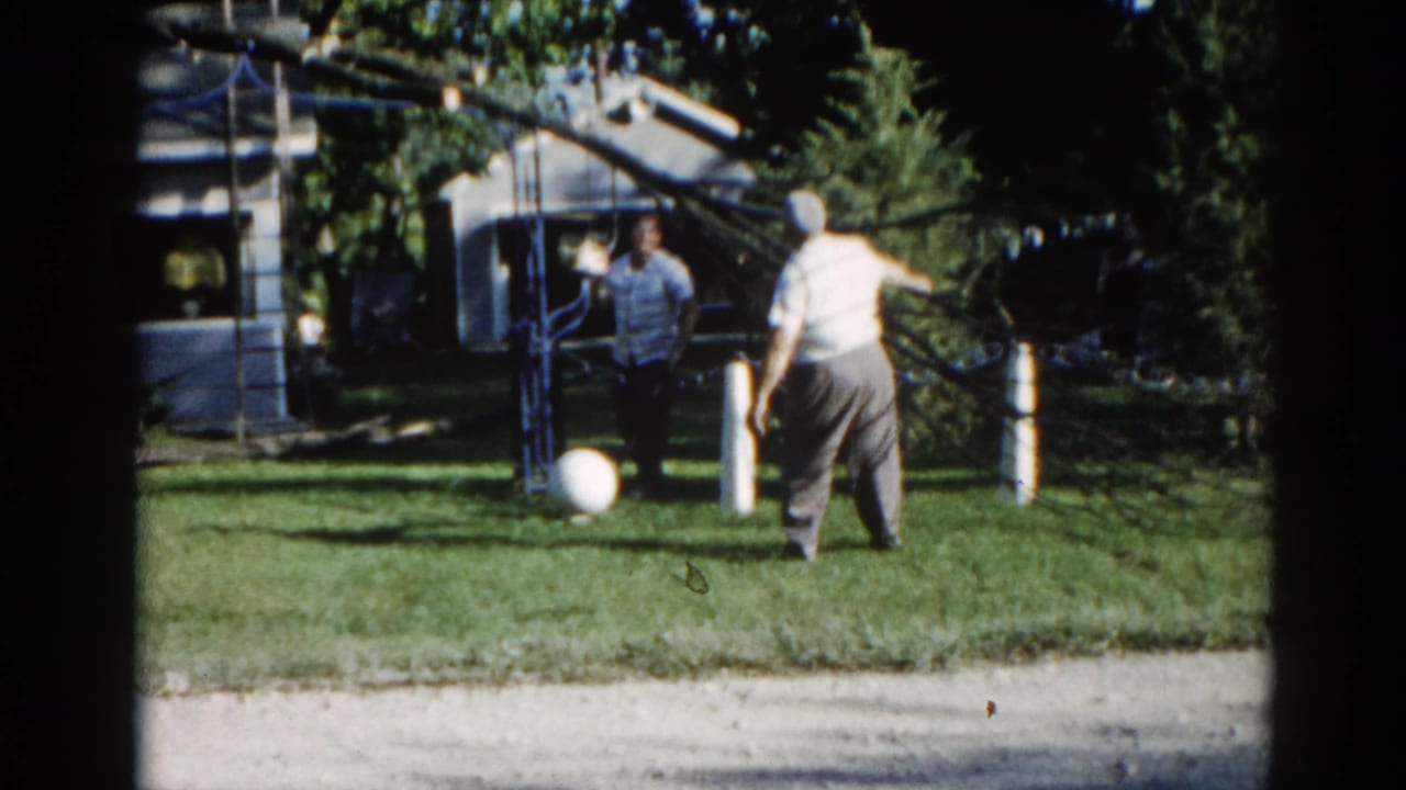 Two adults in a suburban backyard, Illinois, circa 1962; man in light shirt facing a metal overhead structure, companion in darker clothing, white ball on grass.