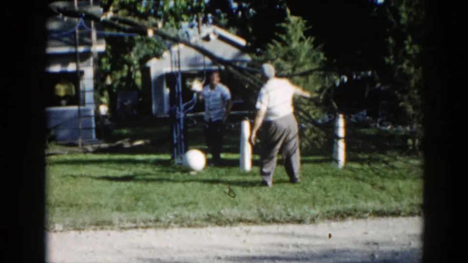 Two adults in a suburban backyard, Illinois, circa 1962; man in light shirt facing a metal overhead structure, companion in darker clothing, white ball on grass.