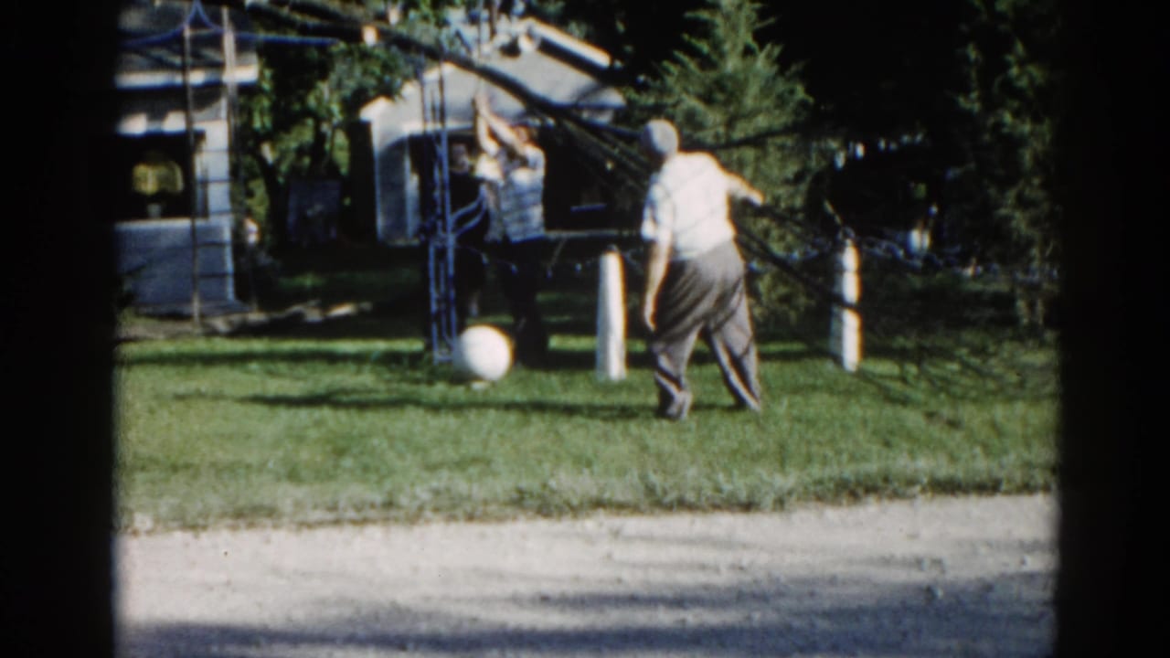 Man in light shirt reaches upward toward an overhead yard structure; second figure stands nearby; white ball rests on the lawn.