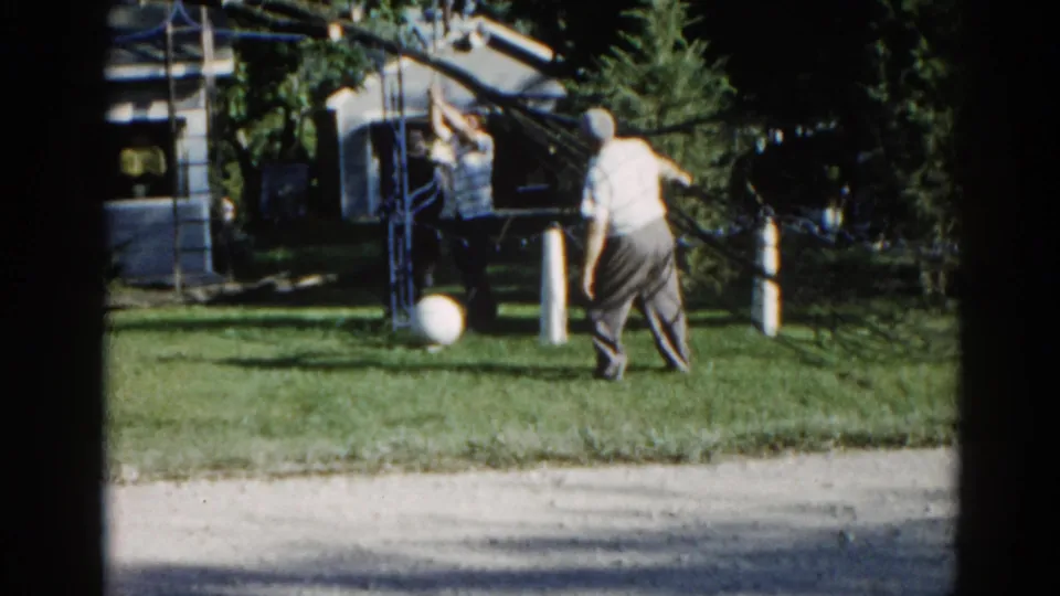 Man in light shirt and dark trousers reaching upward toward a metal yard structure in a suburban Illinois backyard, circa 1962; companion and white ball visible.