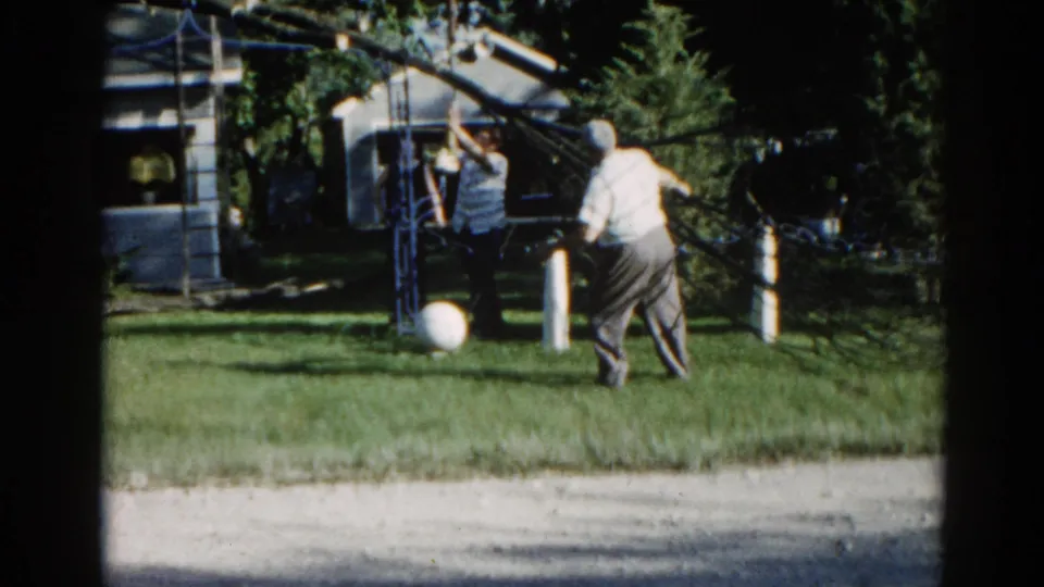 Two adults near an overhead yard structure in a sunlit suburban backyard, Illinois, circa 1962; white ball on lawn, modest outbuilding behind.