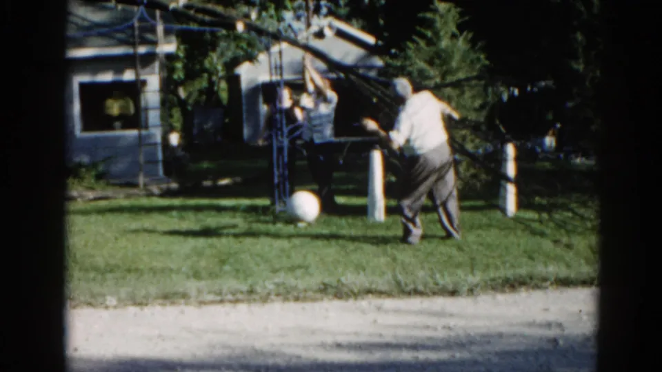 Man standing near an overhead yard structure in a suburban Illinois backyard, circa 1962; companion visible, white ball on green grass, small outbuilding behind.