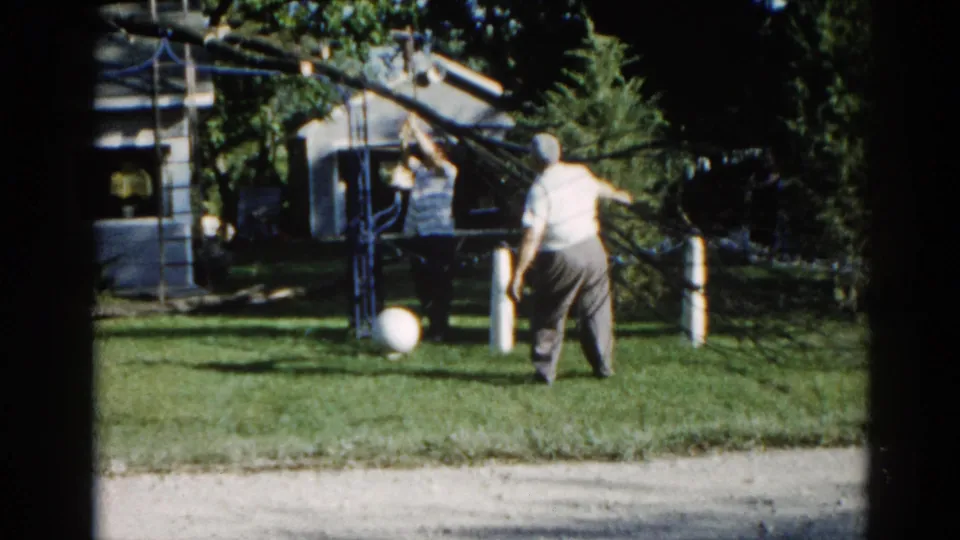 Two figures working in a suburban Illinois yard, c. 1962; fallen tree branch overhead, ranch house in background, second person near fence visible, shot on 8mm.